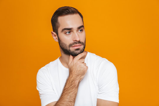 Image of attractive man 30s in t-shirt touching his chin while standing, isolated over yellow background