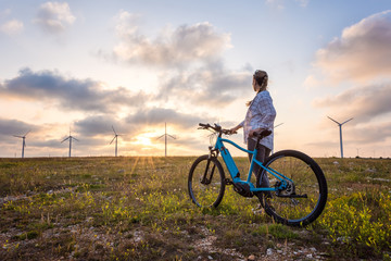 Woman with a bike in the nature / A woman with a bike enjoys the view of sunset over a summer field with a wind farm - obrazy, fototapety, plakaty