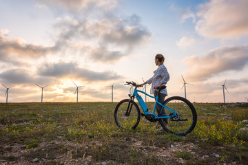 Woman with a bike in the nature / A woman with a bike enjoys the view of sunset over a summer field with a wind farm - obrazy, fototapety, plakaty