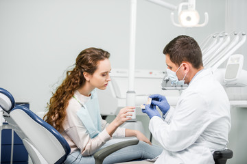 dentist in latex gloves and mask holding teeth model near woman