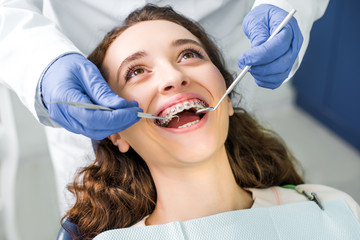 cropped view of dentist in latex gloves examining cheerful woman in braces with opened mouth - obrazy, fototapety, plakaty