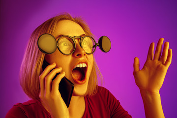Portrait of surprised confused woman looking at mobile phone isolated over pink neon background