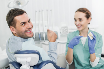 Obraz premium selective focus of handsome man showing thumb up near beautiful dentist holding teeth model