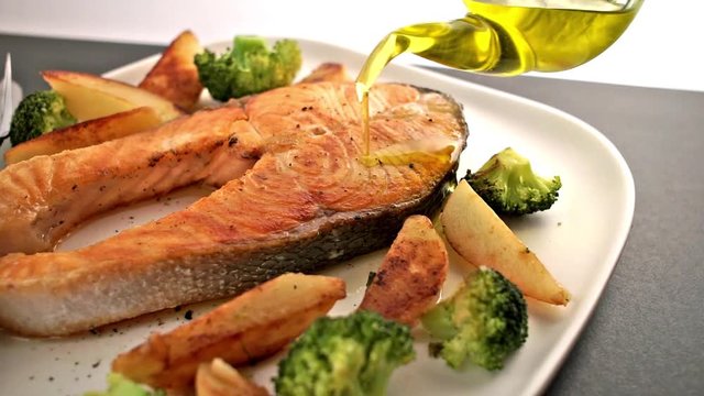Meal with salmon fish steak and vegetable. Bottle of Olive oil pouring on salmon fish. Healthy food. Main course Close up. Slow motion video footage. Slowmo. Slow-mo