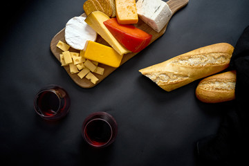 Wooden board with various types of cheese and baguette and two glasses of red wine on black table