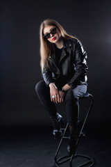 young girl in a black leather jacket and sunglasses on a dark background in the studio sitting