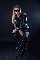 Fototapeta na wymiar young girl in a black leather jacket and sunglasses on a dark background in the studio sitting
