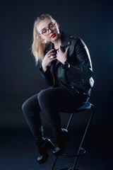 Fototapeta na wymiar young girl in a black leather jacket and glasses on a dark background in the studio sitting