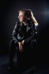 young girl in a black leather jacket and glasses on a dark background in the studio sitting