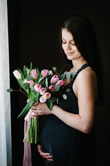 Pregnant woman hold bouquet of flowers in hands, girl embraces a round stomach belly near the window at home. Standing sideways. Maternity concept. Close up. nine months. Baby Shower.