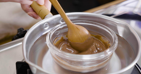 close up focus on hand holding cooking pot with chocolate melting being removed by chef hand on stove of kitchen to make sweet desserts. wooden spoon stirring in creamy cocoa in boiled hot water.