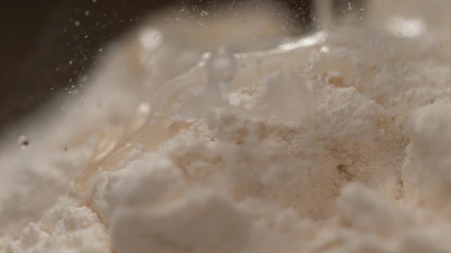SLOW MOTION, MACRO, DOF: Transparent liquid flowing down a heap of clumpy flour in a bakery. Small white particles fly in the air as the shiny viscose liquid flows down a heap of bleached flour.