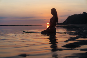 Fototapeta premium Silhouette and reflection of girl sitting on surfboard at ocean beach on background of beautiful sunset