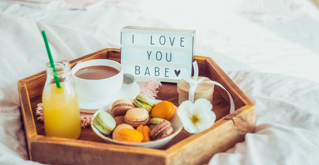 Romantic Breakfast in bed with I love you baby text on lighted box. Cup of coffee, juice, macaroons, flower and gift box on wooden tray. Birthday, Valentine's day morning. Wide banner. Copy space.