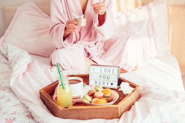 Fototapeta na wymiar Romantic Breakfast in bed with I love you text on lighted box, coffee, macaroons on wooden tray and blurred cropped woman in a bathrobe opening gift box. Birthday, Valentine's day morning. Copy space.