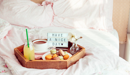 Fototapeta na wymiar Breakfast in bed with Have a nice day text on lighted box. Coffee cup, juice, macaroons, flower in vase on wooden tray. Good morning mood. Hospitality, care, service concept. Wide banner. Copy space.