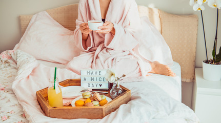 Obraz na płótnie Canvas Good morning mood. Young woman in bathrobe sitting on the bed, drinking coffee and has her breakfast in bed with Have a nice day text on lighted box. Hospitality, care, service concept. Copy space.