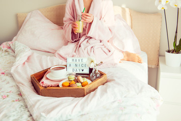 Fototapeta na wymiar Good morning concept. Breakfast in bed with Have a nice day text on lighted box, coffee and macaroons on tray and woman in bathrobe drinking juice. Hospitality, care, service. Square. Copy space.