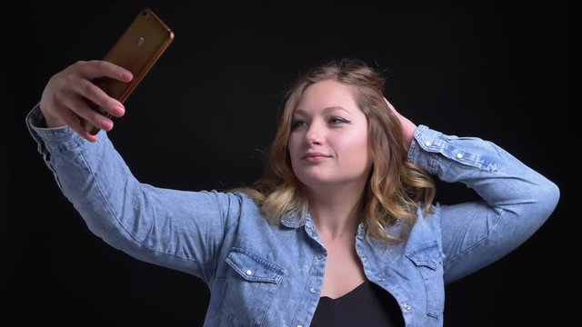 Closeup portrait of adult caucasian blonde female taking selfies on the phone in front of the camera with background isolated on black