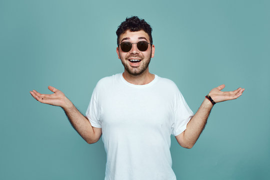 I don't now. Bearded man in sunglasses and white t sirt is gesturing unknown sign isolated over blue background. Young hipster with confuce face expression
