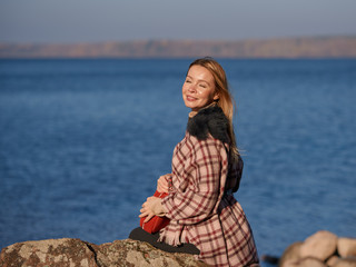 Fototapeta na wymiar Beautiful European woman with long blond hair is sitting on the big stone on the bank of the lake. She is enjoying the serenity around, her eyes are closed and she is smiling.