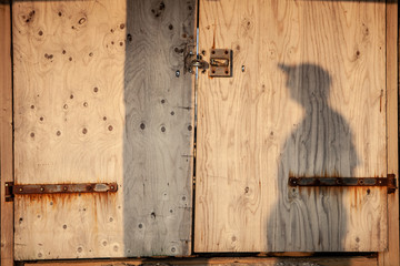 Alone shadow of a man with a hat on a wooden closed door with rusty metal elements. Warm atmosphere.