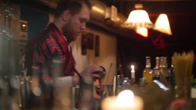 Young professional bartender pouring red drink into the glass in the bar with soft interior lighting.