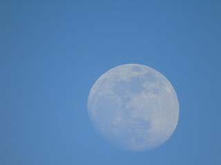 Liguria, Italy - 02/18/2019: An amazing photography of the full moonlight over the mountains, hidden and out of the trees in the village by day with beautiful blue sky in the background in winter days