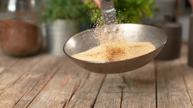SLOW MOTION, MACRO, DOF: Breadcrumbs fall on the table as the female chef mixes them in a hot pan. Unrecognizable woman swirling stale breadcrumbs in a metal pan over a rustic wooden kitchen counter.