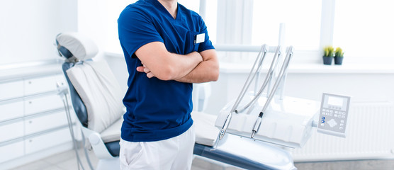 Male dentist in blue in bright white room standing with his hands crossed. no head copyspace