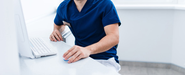 Female dentist doctor in blue sitting at workplace witc pc computer in bright room. no head copyspace
