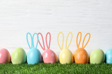 Set of colorful eggs with Easter bunny ears on green grass against wooden background, space for text