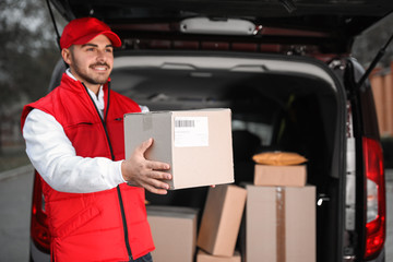 Deliveryman in uniform giving parcel near van outdoors