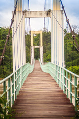 Vintage suspension bridge hanging across the river. Old lengthy hanging wooden footbridge with steel rails over river against cloudy sky background. Hanging wooden bridge at Kaengtana national park.