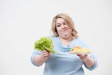 Young fluffy fat woman in casual blue clothes on a white background holding green salad leaves and a plate of fast food, hamburger and fries. Diet and proper nutrition.