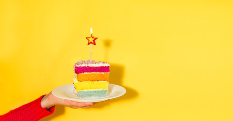 Female hand holding white plate with slice of Rainbow cake with birning candle in the shape of star...