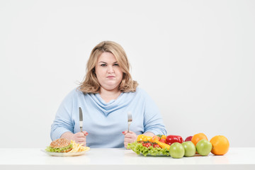 Young curvy fat woman in casual blue clothes on a white background at the table chooses what to eat: fast food or healthy food. Diet and proper nutrition.