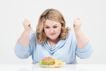 Young curvy fat woman in casual blue clothes on a white background at the table eagerly eating fast food, hamburger and french fries. Diet and proper nutrition.