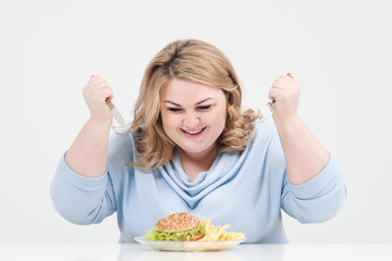 Young curvy fat woman in casual blue clothes on a white background at the table eagerly eating fast...