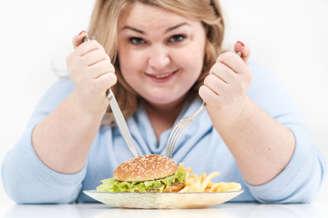 Obraz na płótnie Canvas Young curvy fat woman in casual blue clothes on a white background at the table eagerly eating fast food, hamburger and french fries. Diet and proper nutrition.