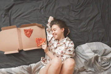 Inside the portrait of a happy girl in pajamas, she lays on a bed with a box of pizza and holds a...