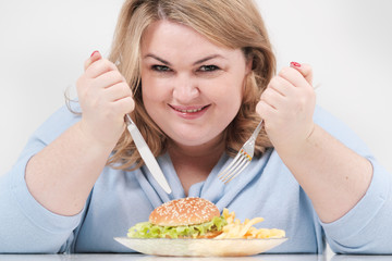 Young curvy fat woman in casual blue clothes on a white background at the table eagerly eating fast...