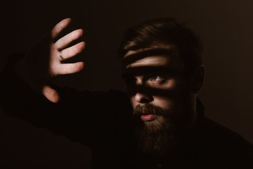 Sepia portrait of a stylish man with a beard and stylish hairdo dressed in the black shirt holds his hand in front of his face on the dark background