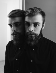 Obraz premium Black and white photo of a man with a beard and stylish hairdo dressed in the black shirt standing next to the mirror with his reflection