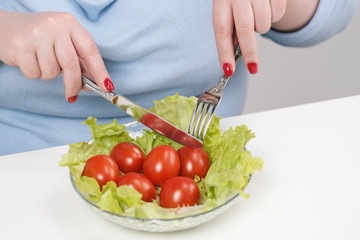 Young lush fat woman in casual blue clothes on a white background at the table and eats a vegetable salad with tomatoes. Diet and proper nutrition.