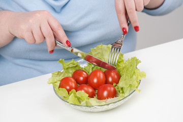 Young lush fat woman in casual blue clothes on a white background at the table and eats a vegetable salad with tomatoes. Diet and proper nutrition.