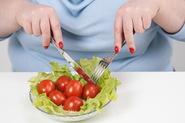 Obraz na płótnie Canvas Young lush fat woman in casual blue clothes on a white background at the table and eats a vegetable salad with tomatoes. Diet and proper nutrition.