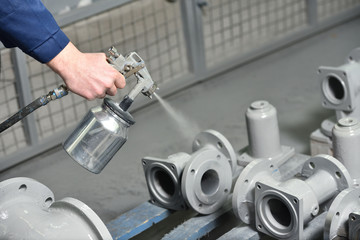 A worker at a factory in a special room paints parts of the valves from the spray gun. Paint valves in gray color, hand and spray gun close up