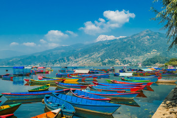Fototapeta na wymiar Many colourful boats parked on the shore of Phewa lake, Pokhara, Nepal. In the back Himalayan mountain range. Small villages visible on the shore. Peaceful and chilled atmosphere. Place to relax.