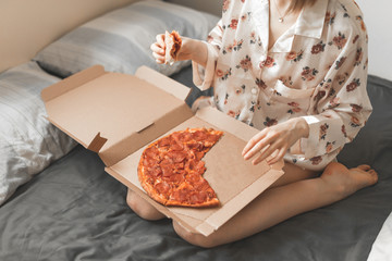 Obraz na płótnie Canvas Close up photo, girl in a pajama sitting on a dark sheet, with a box of pizza on her lap, holding a piece of pizza in her hands, eating fast food on the bed. Copyspace